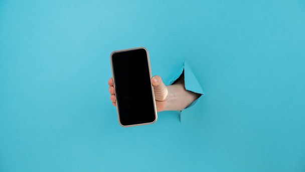 A female hand sticking out of a hole from a blue background holds a smartphone with a black screen - Фото, зображення