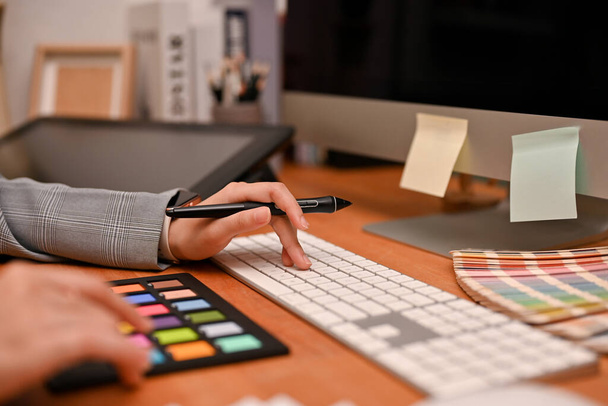 Μια επιτυχημένη γυναίκα γραφίστρια που κρατάει μια γραφίδα, πληκτρολογεί σε πληκτρολόγιο υπολογιστή και χρησιμοποιεί πίνακα ελέγχου χρώματος. περικοπή εικόνας - Φωτογραφία, εικόνα