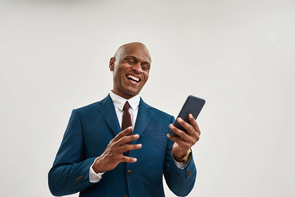 Cheerful african american businessman watching smartphone. Bald adult man wearing suit. Concept of modern successful male lifestyle. Isolated on white background. Studio shoot. Copy space - Photo, image