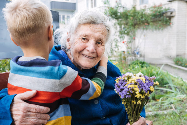 Grandson boy giving a flower to grandma. Grandson and grandmother spending time together. Granny with grandsons enjoying time together outdoor. - Photo, Image