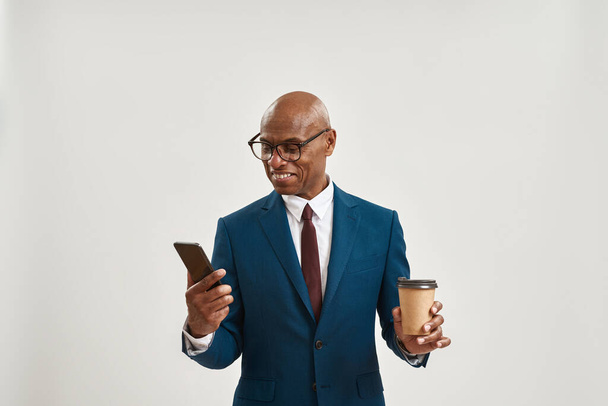 Smiling african american businessman with coffee watching smartphone. Bald adult man wearing suit and glasses. Concept of modern successful male lifestyle. White background. Studio shoot. Copy space - Photo, Image