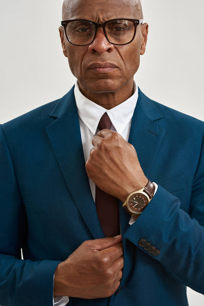 Cropped image and obscure face of serious adult african american company leader wear glasses straighten tie of his suit. Modern successful male lifestyle. Isolated on white background. Studio shoot - Фото, изображение