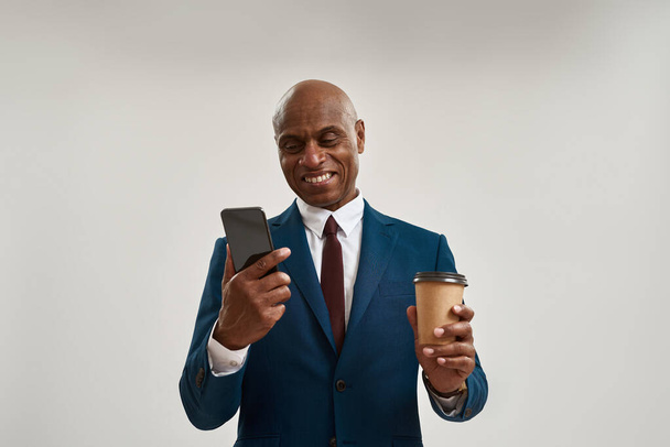 Smiling african american company leader with coffee watching smartphone. Bald adult man wearing suit. Concept of modern successful male lifestyle. Isolated on white background in studio. Copy space - Fotoğraf, Görsel