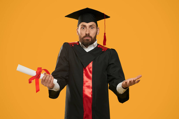 Front view of surprised, shocked student looking at camera. Handsome male with beard holding diploma, graduating from college, university. Isolated on yellow studio background. - Foto, Bild