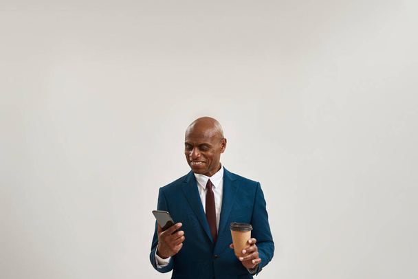 Smiling african american businessman with coffee watching smartphone. Bald adult man wearing suit. Concept of modern successful male lifestyle. Isolated on white background. Studio shoot. Copy space - Fotoğraf, Görsel