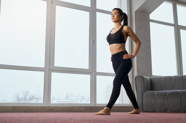 Side view of caucasian woman staying in position of yoga, looking straight while training at home. Slim female yogi with ponytail practicing yoga exercise in spacious studio. Concept of yoga practice. - Photo, Image