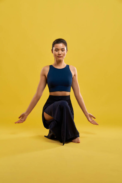 Calm girl meditating in asana, while squatting with hands down indoor. Portrait view of serious lady in black top balancing in yoga pose, isolated on orange studio background. Concept of meditation.  - Photo, Image