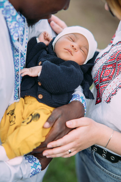 ウクライナの民族刺繍シャツを着た幸せな異人種間の家族は彼らの新生児を保持する。異人種間の家族と異なる人種間の団結の概念。閉鎖. - 写真・画像