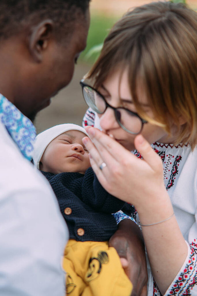 ウクライナの民族刺繍シャツを着て幸せな異人種間の家族は、新生児の手を保持し、キス。異人種間の家族と団結の概念. - 写真・画像