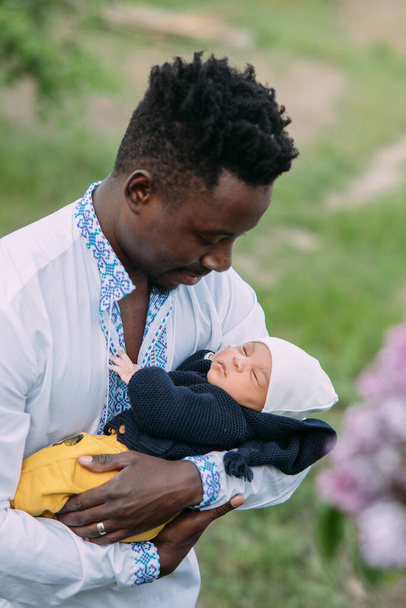 ウクライナの民族刺繍のシャツを着た幸せなアフリカ人男性は、異人種間の結婚から生まれたばかりの赤ちゃんを保持しています。異人種間の家族と団結の概念. - 写真・画像