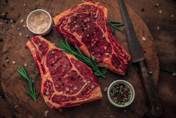 table top of fresh beef steak with sprinkles of salt and pepper and green herbs on top of old wooden chopping board with small bowls of salt and pepper and knife - Foto, Bild