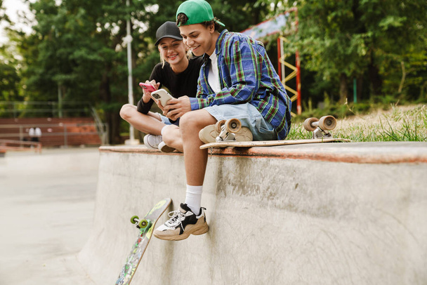 Two multiracial teenagers smiling and using cellphones while spending time at skate park outdoors - Фото, изображение