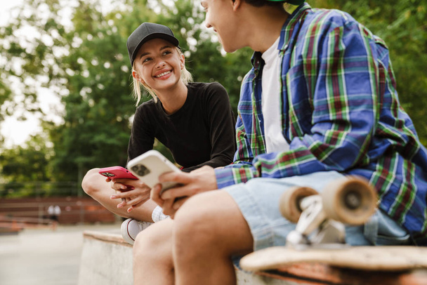 Two multiracial teenagers smiling and using cellphones while spending time at skate park outdoors - Fotó, kép