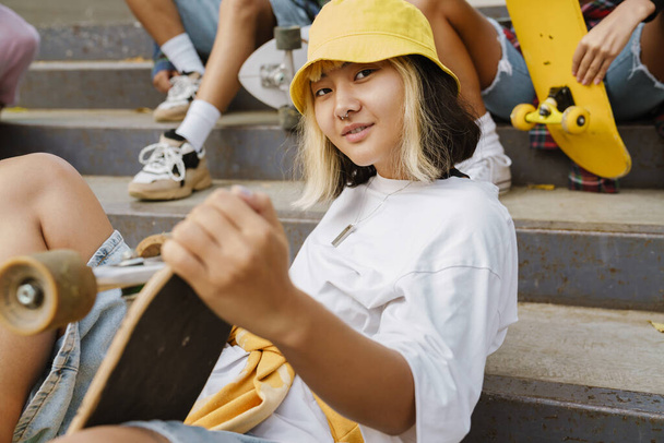 Asian girl sitting on stairs while spending time with her friends at skate park outdoors - Foto, Imagen