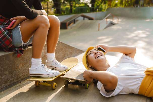 Asian girl laughing and talking on mobile phone while lying at skate park outdoors - Photo, image