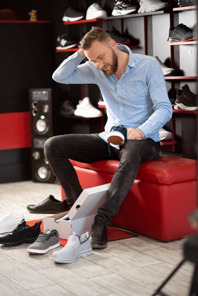 Kaukaski brodaty mężczyzna wybiera buty turystyczne siedząc w pomieszczeniu nowoczesnego sklepu sportowego. Koncepcja złożoności wyboru i duży asortyment. przestrzeń kopiowania. - Zdjęcie, obraz