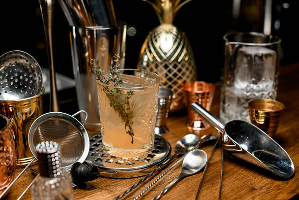close-up of a transparent crystal glass with alcoholic cocktail decorated with a branch of greenery on a bar counter surrounded by various bar accessories - Fotó, kép