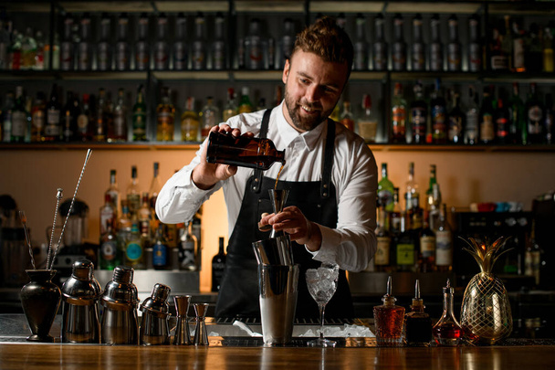 beautiful view of the bar on which there are different shakers and bottles, and the bartender pours a drink from a bottle into a jigger - Zdjęcie, obraz