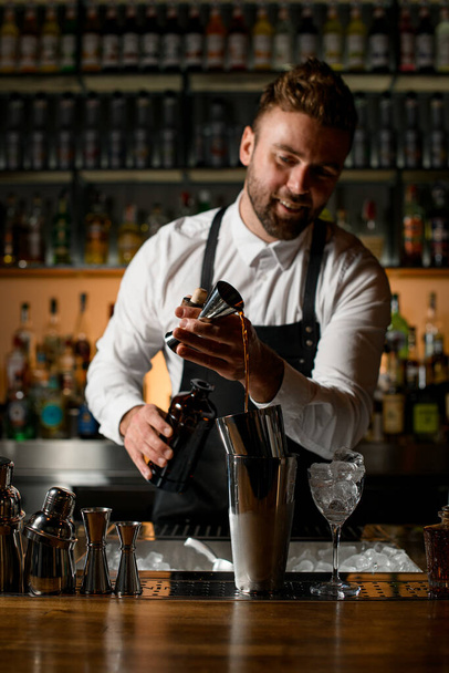 бар счетчик с различными шейкерами и бутылками на нем и мужчина бармен осторожно наливает алкогольный напиток из jigger в шейкер чашку - Фото, изображение