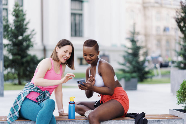 Cheerful smiling friends in sportswear sitting on a bench in the city discussing while using smartphone looking to screen. Multiethnic women having a fitness workout break. - Foto, afbeelding
