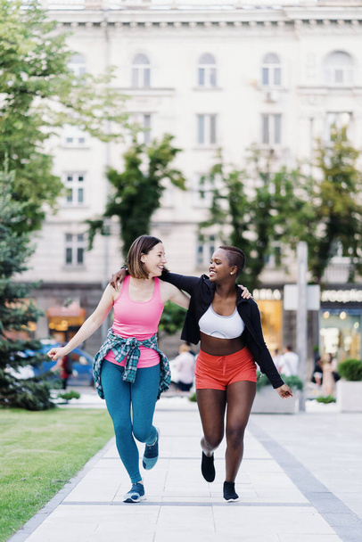 Vrolijke glimlachende vrienden in sportkleding rennen in de stad dicussing. Multi-etnische vrouwen met een fitnesstraining. - Foto, afbeelding