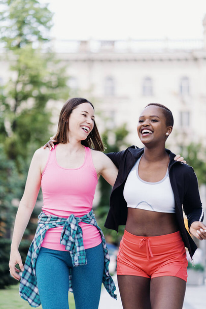 Alegres amigos sonrientes en ropa deportiva caminando después de una sesión deportiva en la ciudad discutiendo y divirtiéndose. Mujeres multiétnicas haciendo ejercicio físico trotando. - Foto, imagen