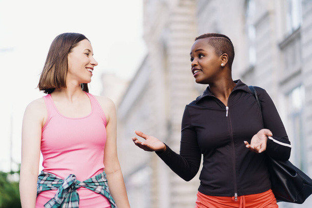 Χαρούμενοι χαμογελαστοί φίλοι με αθλητικά ρούχα περπατούν μετά από μια αθλητική συνεδρία στην πόλη συζητώντας και διασκεδάζοντας. Πολυεθνικές γυναίκες που κάνουν τζόκινγκ γυμναστικής. - Φωτογραφία, εικόνα
