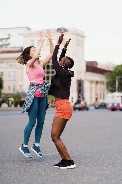 Sonriendo amigos alegres en ropa deportiva corriendo en la ciudad discutiendo, dando hola cinco, celebrando el éxito y la felicidad. Mujeres multiétnicas que hacen ejercicio físico. - Foto, imagen