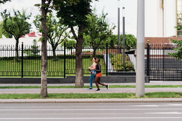 Buenos riends en ropa deportiva corriendo en la ciudad dicussing. Mujeres multiétnicas que hacen ejercicio físico. - Foto, Imagen