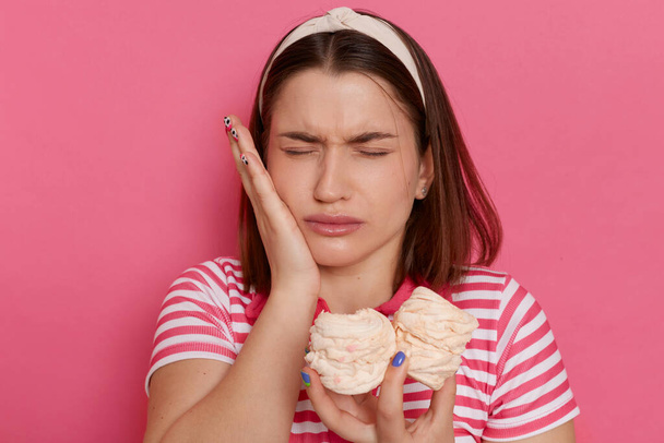 Indoor shot of sad unhealthy woman wearing striped t shirt posing isolated over pink background, feels pain in teeth after eating sugary marshmallows, keeping hand on cheek. - Valokuva, kuva
