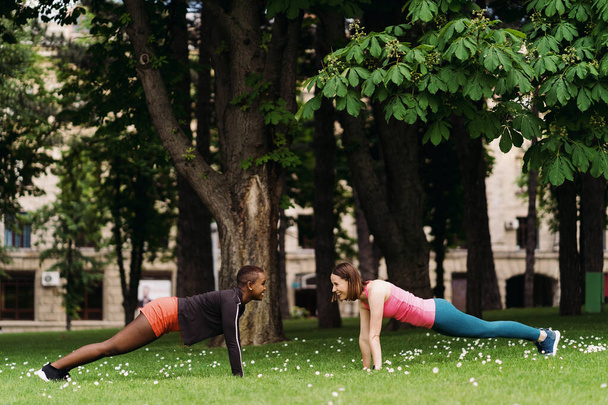Twee lachende divers jonge vrouw in atletische workout kleren doen een plank oefening uit te werken samen in het park op gras. - Foto, afbeelding
