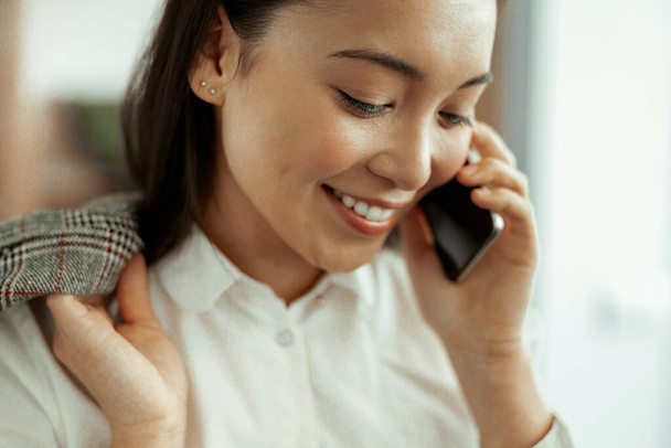 Крупным планом улыбается азиатская деловая женщина, делающая телефонный звонок стоя в офисе. Высокое качество фото - Фото, изображение