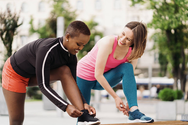 Vrolijke glimlachende vrienden in sportkleding gelijkmakende sportschoenen veters in de stad bespreken. Multi-etnische vrouwen met een fitness workout break. - Foto, afbeelding