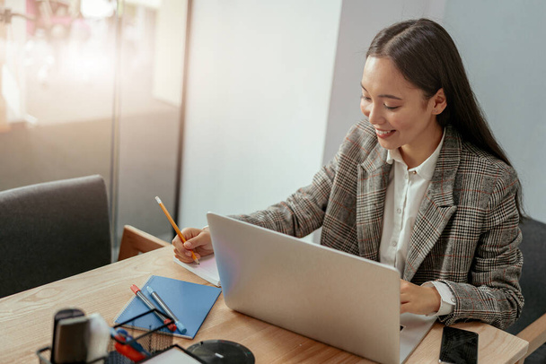 オフィスでノートパソコンを操作しながらノートを作っている女性の笑顔。ぼやけた背景 - 写真・画像