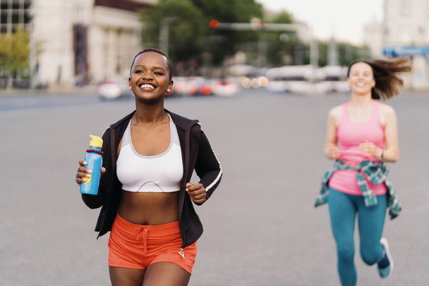 Weseli, uśmiechnięci przyjaciele w sportowej odzieży biegający po mieście i dyskutujący. Wieloetniczne kobiety trenujące fitness. - Zdjęcie, obraz