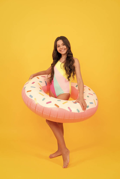 glückliches Strandkind mit lockigem Haar im Badeanzug mit Donut-Aufblasring für Pool-Party-Spaß im Sommerurlaub auf gelbem Hintergrund, Kindheit. - Foto, Bild
