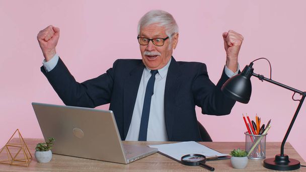 Oh my God Wow. Senior business man, using laptop, analysing paperwork documents raising hands in surprise, shocked by sudden victory, good news. Elderly freelancer working at office pink background - Foto, Imagem