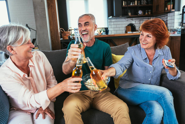 Grupa starszych przyjaciół o popcorn i piwo oglądanie telewizji razem w weekend w domu - Zdjęcie, obraz