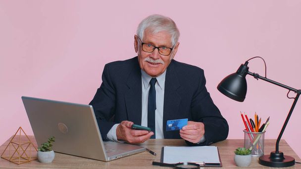 Senior Geschäftsmann mit Kreditkarte und Smartphone während der Überweisung von Geld, Online-Einkäufe sitzt am Schreibtisch im Büro. Älterer Mann isoliert drinnen auf rosa Studiowand Hintergrund - Foto, Bild