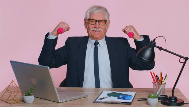 Senior businessman working out, pumping up arm muscles lifting pink dumbbells, relaxing after work, health care at office. Sport motivation for elderly man isolated indoor on pink studio background - Photo, Image