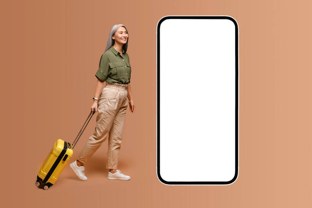 Mobile app for travelers. Senior Asian gray-haired modern retired woman with yellow luggage suitcase walking to huge smartphone with empty display, copy space, mockup - Foto, imagen