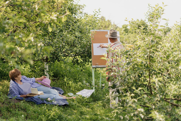 HAPPY SENIOR COUPLE περνούν χρόνο μαζί στον κήπο ή τη φύση τους. Ένας άντρας ζωγραφίζει τη γυναίκα του ενώ αυτή χαλαρώνει με ένα βιβλίο. Μεγαλώνουμε όμορφα μαζί. Ζωντανός πράσινος κήπος - Φωτογραφία, εικόνα