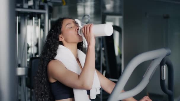 Водный баланс во время спортивных тренировок. Молодая активная спортсменка пьет из бутылки, тренируется на тренажере в современном тренажерном зале, свободное место - Кадры, видео