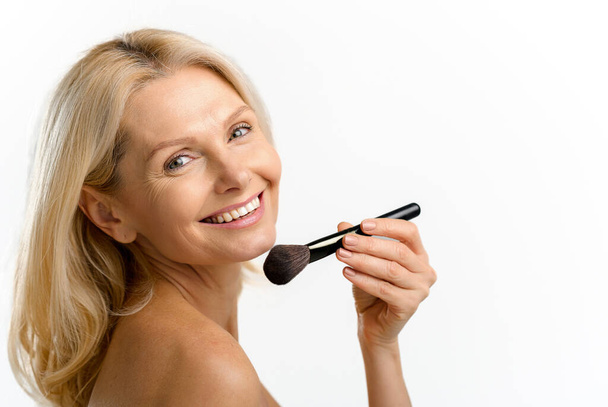 Charming blonde 40s 50s middle aged woman with holding make up brush on shining face with perfect skin, looking at the camera, applying powder or some cosmetic product, advertising concept - Fotó, kép