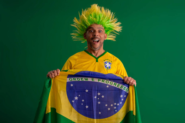 Hombre afrobrasileño, partidario de la selección de fútbol brasileña en la copa 2022, con la bandera de Brasil y con expresiones faciales. - Foto, imagen