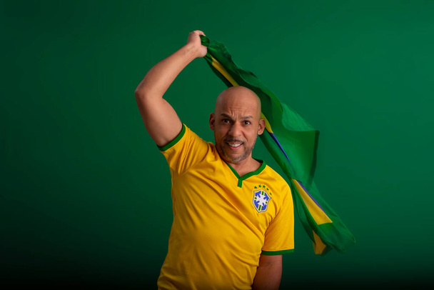 Afro Brazilian man, supporter of the Brazilian football team in the 2022 cup, with the flag of Brazil and with facial expressions. - 写真・画像