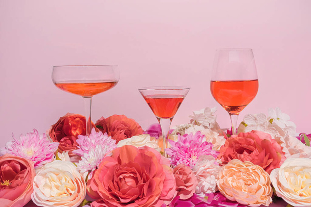 Διασκέδαση, πολύχρωμη καλοκαιρινή σύνθεση με τρία ποτήρια κρασιού και κοκτέιλ και ροζ και κόκκινα λουλούδια. Φωτεινή και μοντέρνα θηλυκή έννοια. - Φωτογραφία, εικόνα