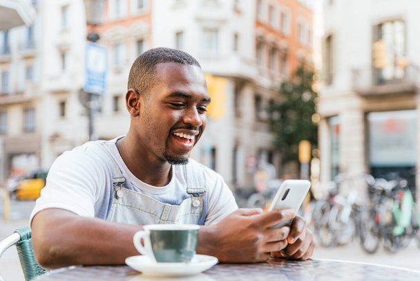 Веселый молодой афроамериканец в повседневной одежде. Он счастливо улыбается, читая хорошие новости на смартфоне, сидя за столом с чашкой кофе в уличном кафе. - Фото, изображение