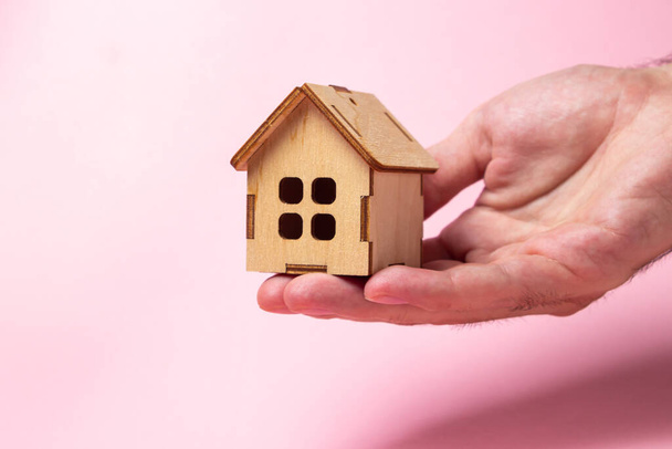 Рука тримає дерев'яний будинок на рожевому тлі. Концепція купівлі-продажу нерухомості. Послуги з нерухомості. Домашнє страхування
 - Фото, зображення
