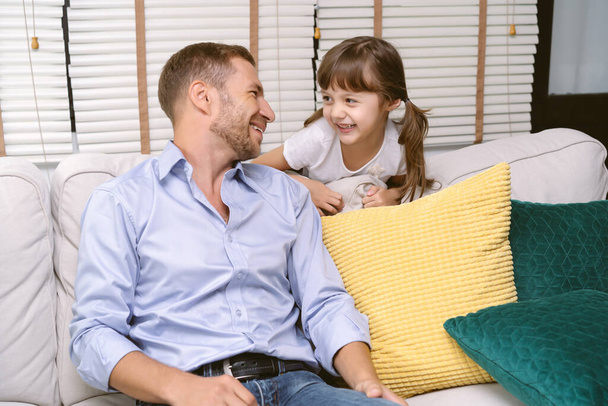 Retrato del padre y su linda hija mirándose mientras el padre está sentado en el sofá y la niña detrás del sofá en la sala de estar en casa. Padre e hija felices pasan tiempo juntos. - Foto, Imagen